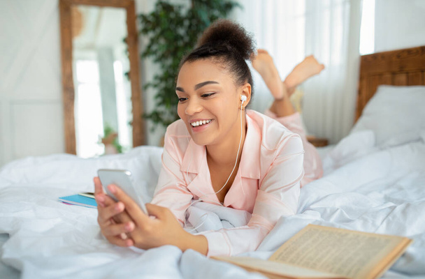 Jonge Afrikaans-Amerikaanse dame in oortelefoons luisteren naar muziek of het hebben van video-oproep op de smartphone, liggend op bed, kopieer ruimte - Foto, afbeelding