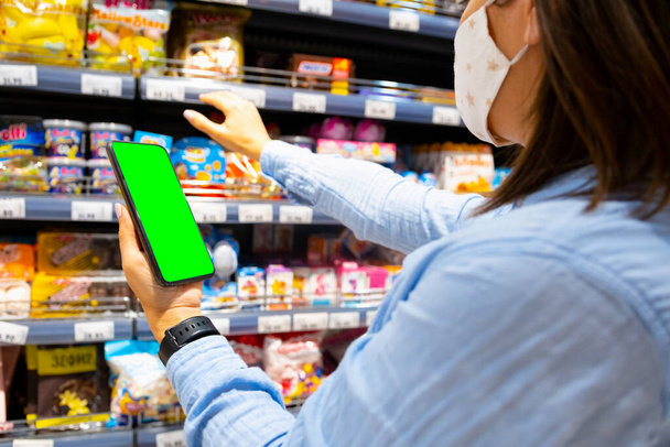 Foto recortada de una joven con máscara facial usando teléfono móvil y comprando comestibles en el supermercado durante la pandemia de virus. - Foto, Imagen