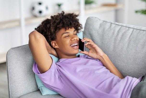 Alegre adolescente negro acostado en el sofá, teniendo una conversación de teléfono inteligente con un amigo en casa - Foto, imagen