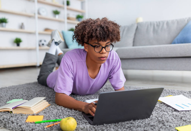 Ukierunkowany czarny nastolatek korzystający z laptopa do zdalnej edukacji, leżący na podłodze i piszący na klawiaturze w domu - Zdjęcie, obraz
