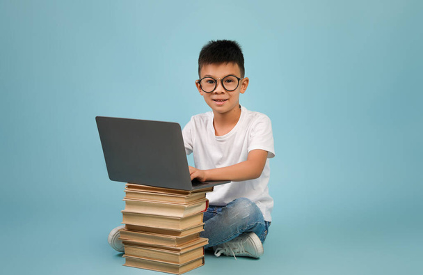 Маленький азиатский мальчик в очках сидит с ноутбуком и стопкой книг - Фото, изображение