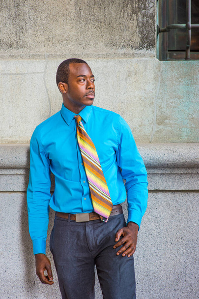 Világoskék ingbe öltözve, mintás nyakkendőben, szürke nadrágban, egy fekete üzletember áll a mintafalnál ablakkal, magabiztosan várva - Fotó, kép