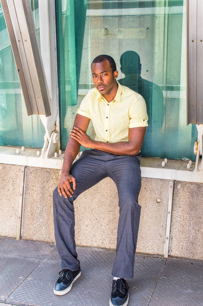 Vestido com uma camisa amarela clara, calças cinza, tênis, um jovem negro atraente está sentado por uma estrutura de metal e contra uma parede de vidro, olhando atenciosamente para você - Foto, Imagem