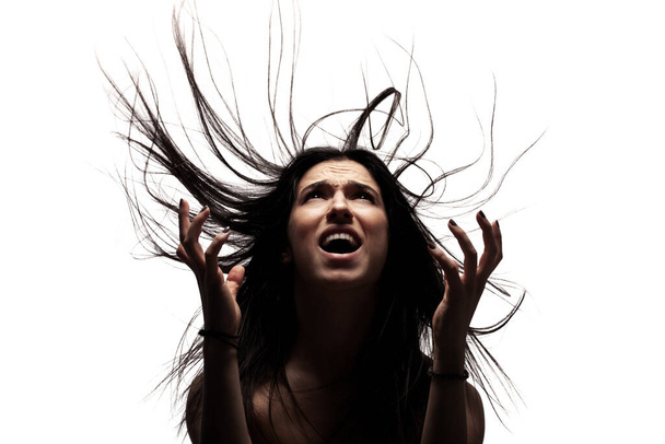 Темный портрет с тенями сердитой девушки с волосами в воздухе, смотрящей вверх и делающей жесты руками. - Фото, изображение