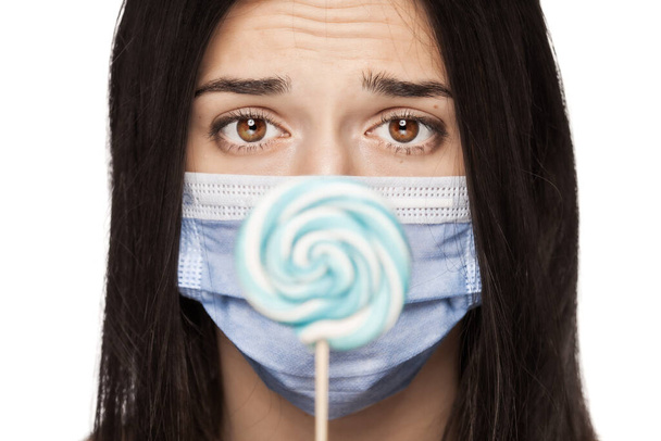 Sad girl having her mouth covered with with medical face mask holding lollipop - Zdjęcie, obraz