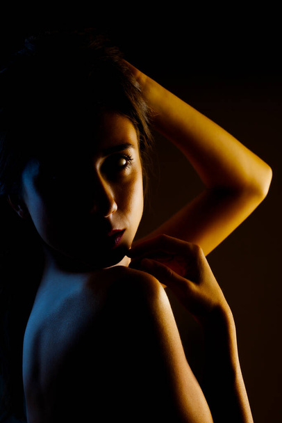 Beautiful brunette girl studio portrait. Side lit half silhouette on dark background. Serious face expression. - 写真・画像