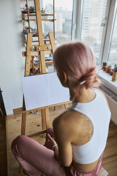 Woman with bun on head sits by easel on light balcony at home - Photo, Image