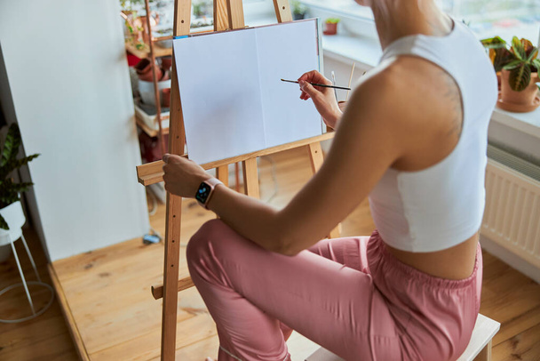 Mujer joven dibuja cuadro sobre lienzo con pincel en caballete de madera en balcón - Foto, Imagen