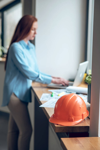 Protective helmet on windowsill and woman working on laptop - Photo, Image