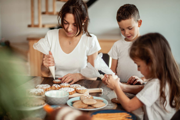 Mom and children decorate Christmas gingerbread at home. A boy and a girl paint with cornets with sugar icing on cookies. New Years decor, branches of a Christmas tree. - Fotografie, Obrázek