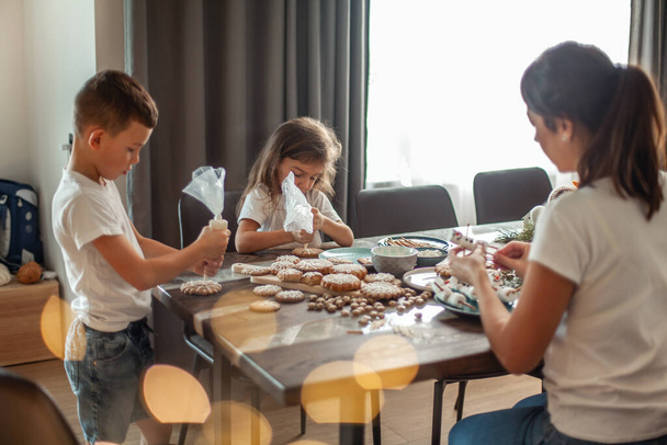 Mom and children decorate Christmas gingerbread at home. A boy and a girl paint with cornets with sugar icing on cookies. New Years decor, branches of a Christmas tree. - Photo, image