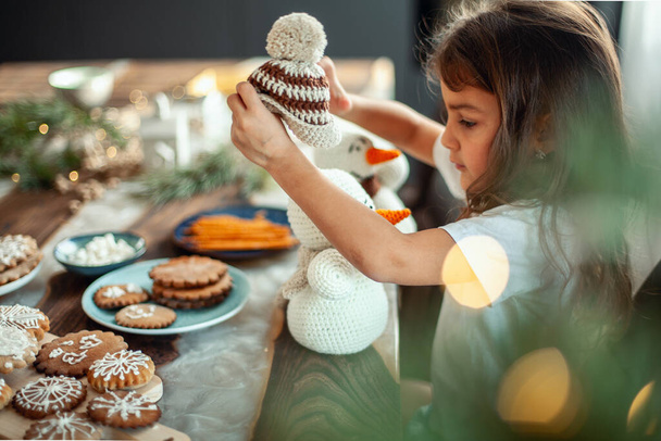 Little cute girl sitting at a table filled with Christmas decor. And he holds two knitted snowmen in his hands. - Foto, immagini