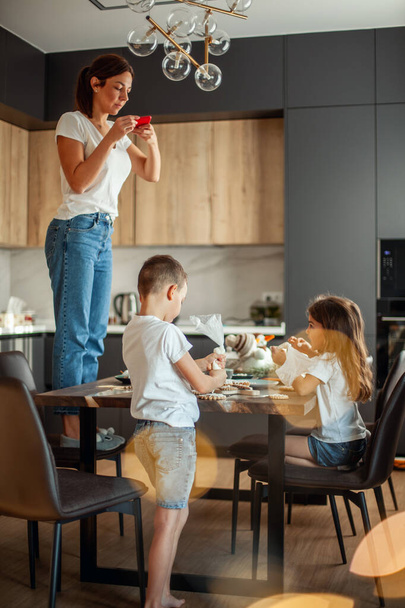 Mom takes pictures of topview as daughter and son decorate gingerbread with sugar icing. Stylish home dining and kitchen. - Φωτογραφία, εικόνα
