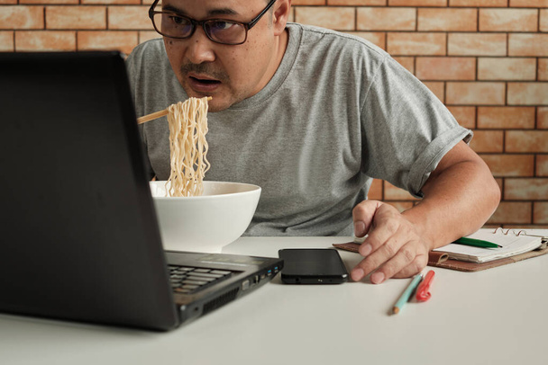 Tajski pracownik męski zajęty pracą z laptopem, używaj pałeczek do szybkiego jedzenia makaronu podczas przerwy na lunch w biurze, ponieważ szybki, smaczny i tani. Z biegiem czasu azjatyckie fast food, niezdrowy styl życia. - Zdjęcie, obraz