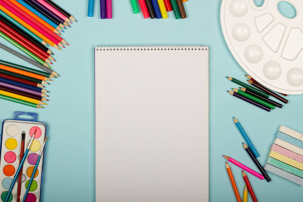 Art school supplies for painting and a blank sheet of paper math notebook on a blue background with copy space for text. Colorful pencils, markers, paints, crayons. Top view. Back to school - Foto, imagen