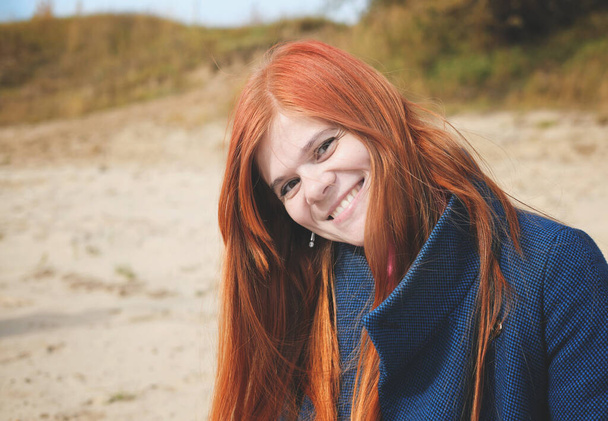 jovem sorrindo mulher de cabelos vermelhos vestindo um casaco azul e olhando para o espectador. Outono retrato de mulher de cabelo de gengibre feliz - Foto, Imagem