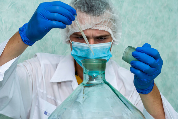 лаборант в белом халате, защитной маске, перчатках и колпачке в лаборатории капает жидкость в сосуд с пипеткой, лицо доктора размыто. Высокое качество фото - Фото, изображение