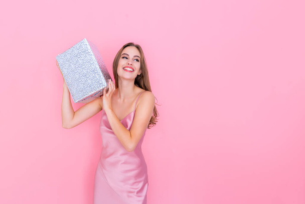 Image of happy girl shaking box with gift wonder what inside wrapped box stands on pink background - Foto, Imagem