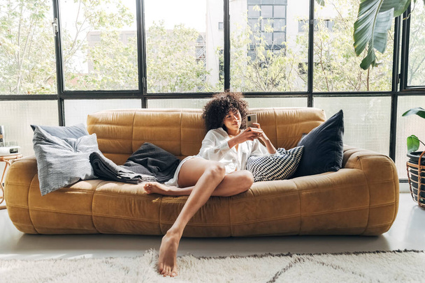 Jolie jeune femme afro-américaine avec les cheveux bouclés assis sur un canapé confortable textos à un ami - Photo, image