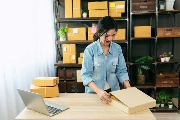 Kadın paket kutusunu masaya koydu. Müşteriye mal teslimatını kontrol ediyor. Asyalı bir kadın ofis masasında küçük bir iş kuruyor. Girişimci Asyalı kadın teslimat için ürün topluyor - Fotoğraf, Görsel