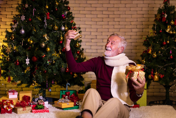 Senior caucasian man celebrating Christmas alone in happiness and excitement while holding his present at nursing home with christmas tree - Foto, Imagen