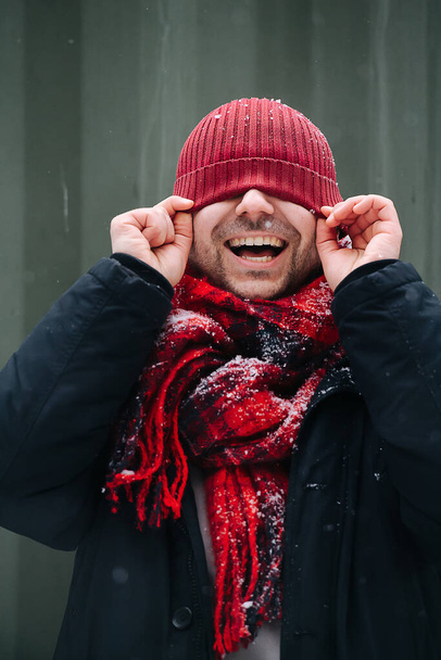 Silly portrait of a positive middle age man with winter hat over his eyes. He wears vibrant red hat and scarf. Smiling openly with teeth. - Фото, изображение