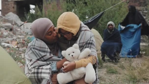 Medium shot of African-American mother and her 8-year-old daughter in warm clothing sitting outdoors under blanket embracing living at refugee camp with other immigrants - Footage, Video
