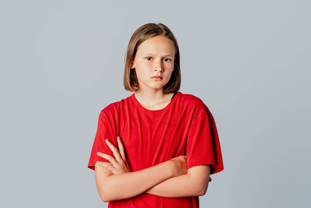 Серьёзно выглядящая оскорбленная девочка-подросток в непринужденной красной футболке, хмурая хмурая от расстройства, неудовлетворенная поза с крестиком на груди, мальчик оскорбил её ожиданием извинений, жалуясь на недовольство, стоять на сером фоне - Фото, изображение