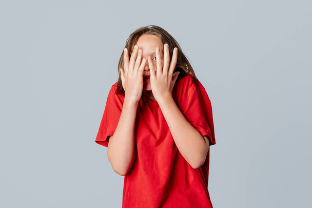 美しいブルネットの10代の女の子は、灰色の背景の上に立って、カジュアルな赤いTシャツを着て、怖い手をカバー顔を怖がってショックを受けた。若い感情的な女性 - 写真・画像