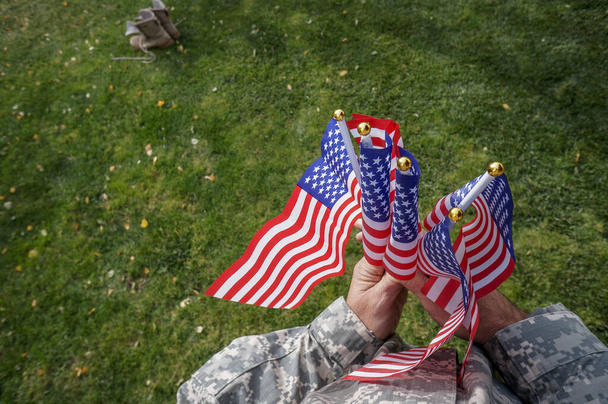 Soldato americano tiene con due mani bandiere che sventolano nel vento su erba verde in un parco in vacanza - Foto, immagini