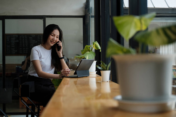 Mujer joven hablando por teléfono móvil y escribiendo notas en una tableta digital mientras está sentada en su escritorio. Asiática mujer trabajando en casa oficina - Foto, imagen