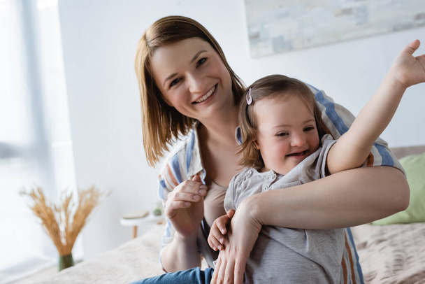 Smiling woman looking at camera while hugging daughter with down syndrome on bed  - Foto, afbeelding