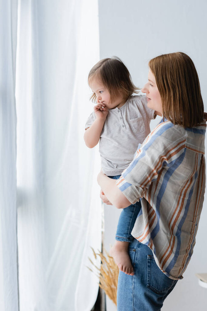 Mujer sosteniendo hija con síndrome de Down cerca de cortinas en casa  - Foto, Imagen