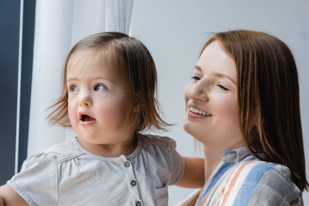 Cheerful woman on blurred background looking at daughter with down syndrome at home  - Foto, afbeelding