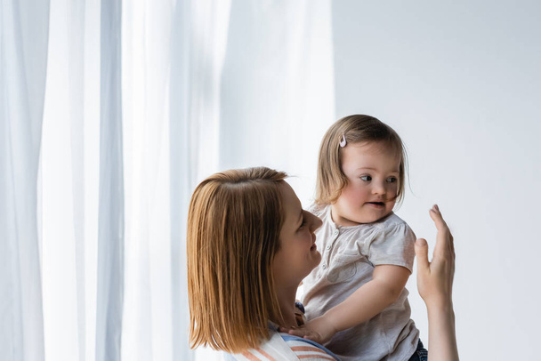 Mujer sonriente mirando a un niño con síndrome de Down cerca de las cortinas en casa  - Foto, Imagen