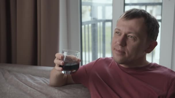 El hombre sueña sentado en casa, sosteniendo un vaso con una bebida en la mano - Imágenes, Vídeo