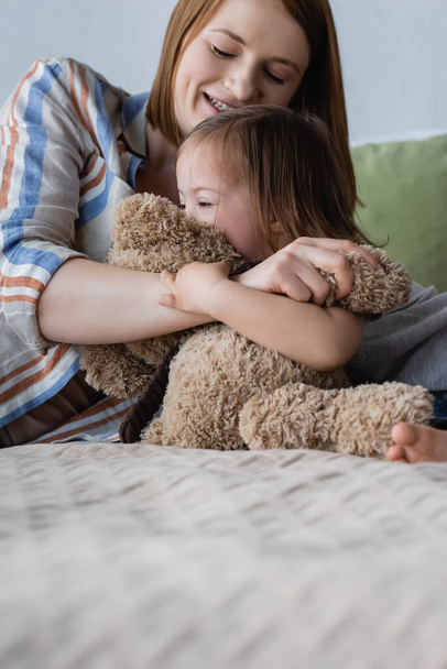 Smiling woman hugging daughter with down syndrome and soft toy on bed  - Photo, Image