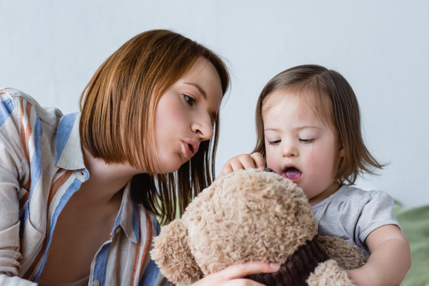 Mother talking to toddler daughter with down syndrome holding soft toy at home  - Valokuva, kuva