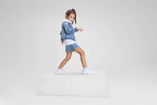Full-length portrait of smiling girl in casual clothes standing on big box isolated on white studio background. Happy childhood concept. - Foto, afbeelding