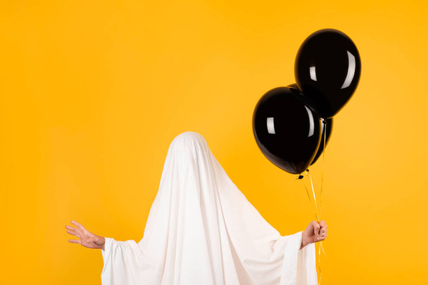 Halloween concept. Girl in a funny white ghost costume holding black balloons in hand. - Photo, Image