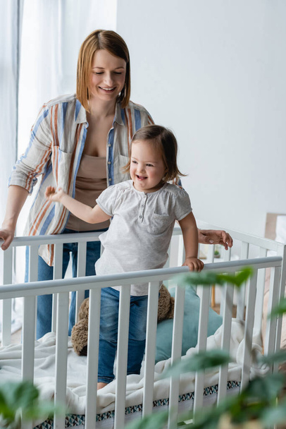 Smiling woman standing near daughter with down syndrome in baby crib  - Φωτογραφία, εικόνα