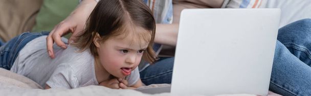 Kid with down syndrome looking at laptop near parent on bed, banner  - Φωτογραφία, εικόνα