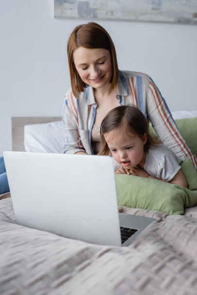 Smiling parent freelancer looking at daughter with down syndrome near laptop on bed  - Φωτογραφία, εικόνα