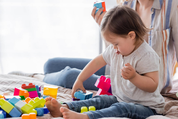Child with down syndrome sticking out tongue while playing building blocks near mother on bed   - 写真・画像