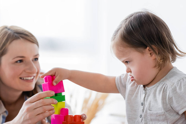 Concentrated kid with down syndrome playing building blocks near blurred smiling mother at home  - Фото, зображення