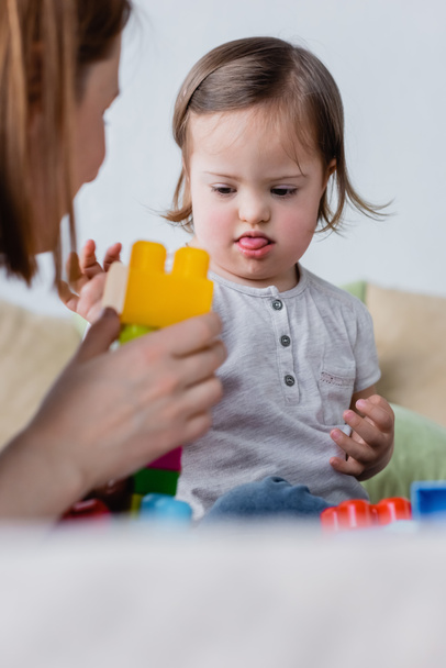 Kid with down syndrome sticking out tongue while playing building blocks with mom at home  - Φωτογραφία, εικόνα
