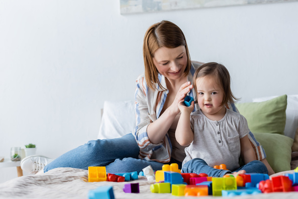 Smiling parent holding building block near daughter with down syndrome on bed  - Foto, afbeelding