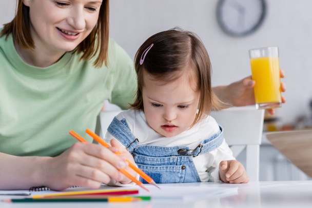 Criança com síndrome de down desenho em papel perto de mãe sorridente com suco de laranja na cozinha  - Foto, Imagem