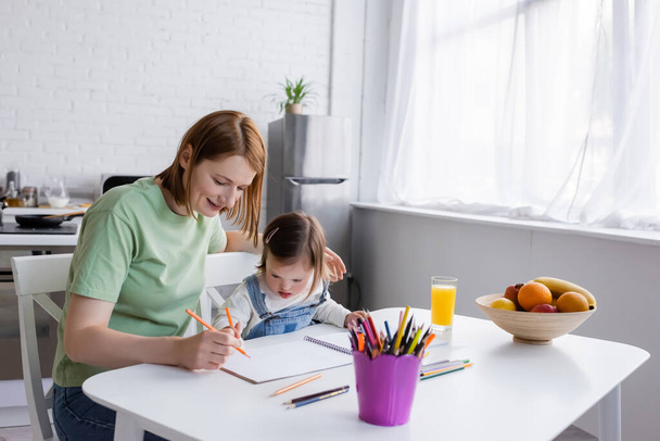 Child with down syndrome drawing on paper near mother and fruits in kitchen  - Photo, Image