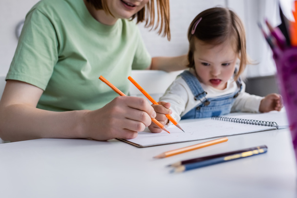 Blurred kid with down syndrome drawing on paper near mom in kitchen  - Photo, Image